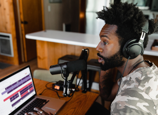 Ein junger Mann sitzt vor einem Laptop und Mikrofon und nimmt einen Podcast auf.